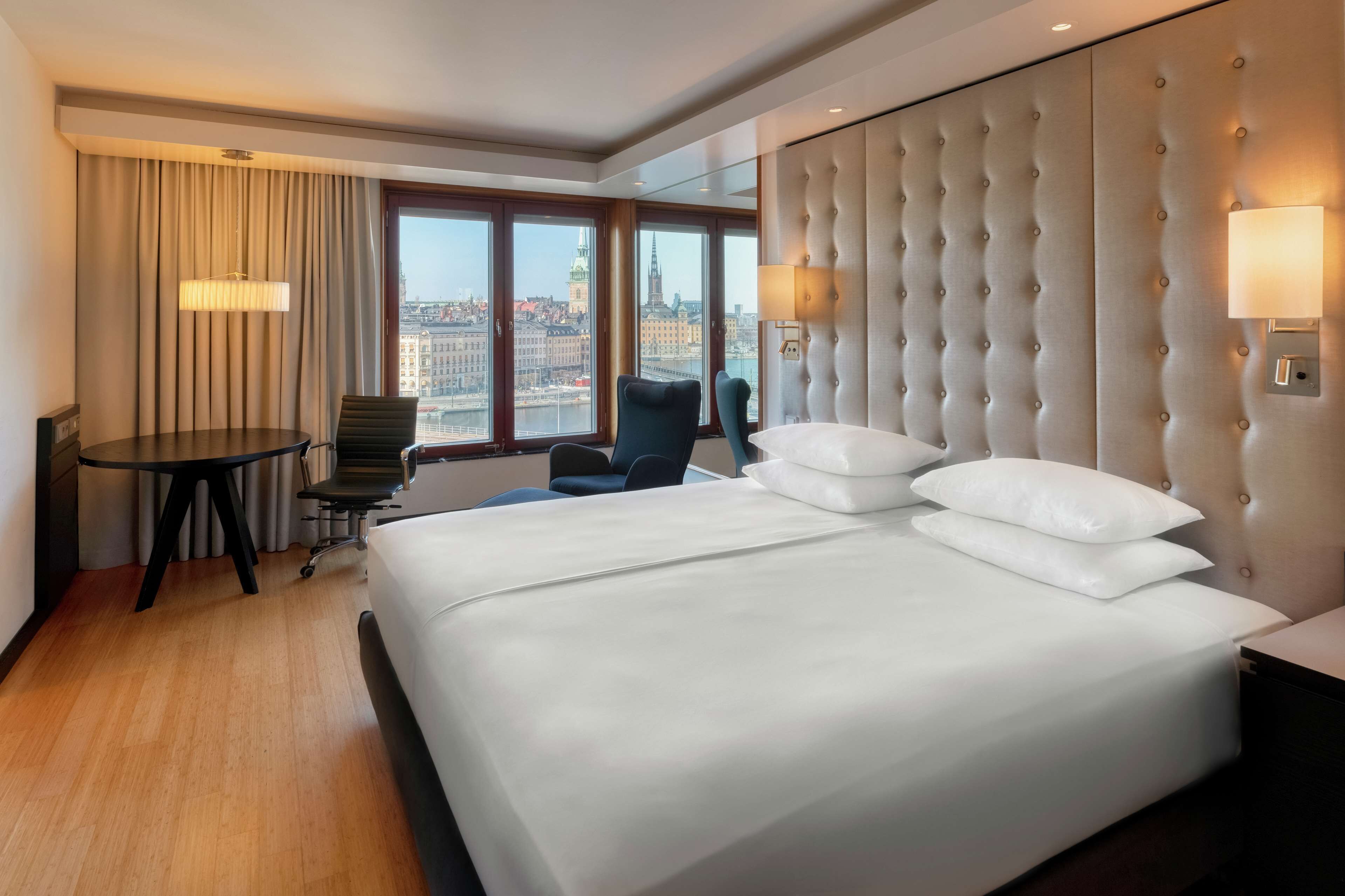 A hotel room with a large bed, a desk, a chair, and a view of a cityscape.