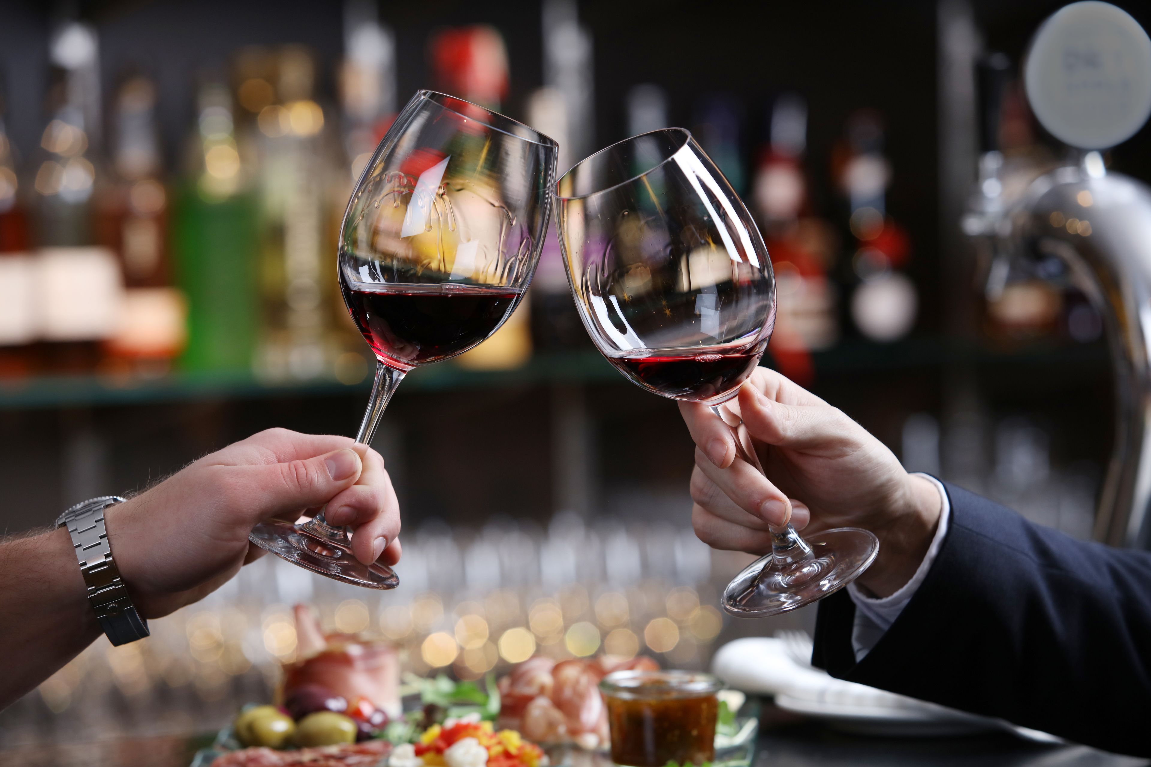 Two people toasting with wine glasses at a bar with bottles in the background.