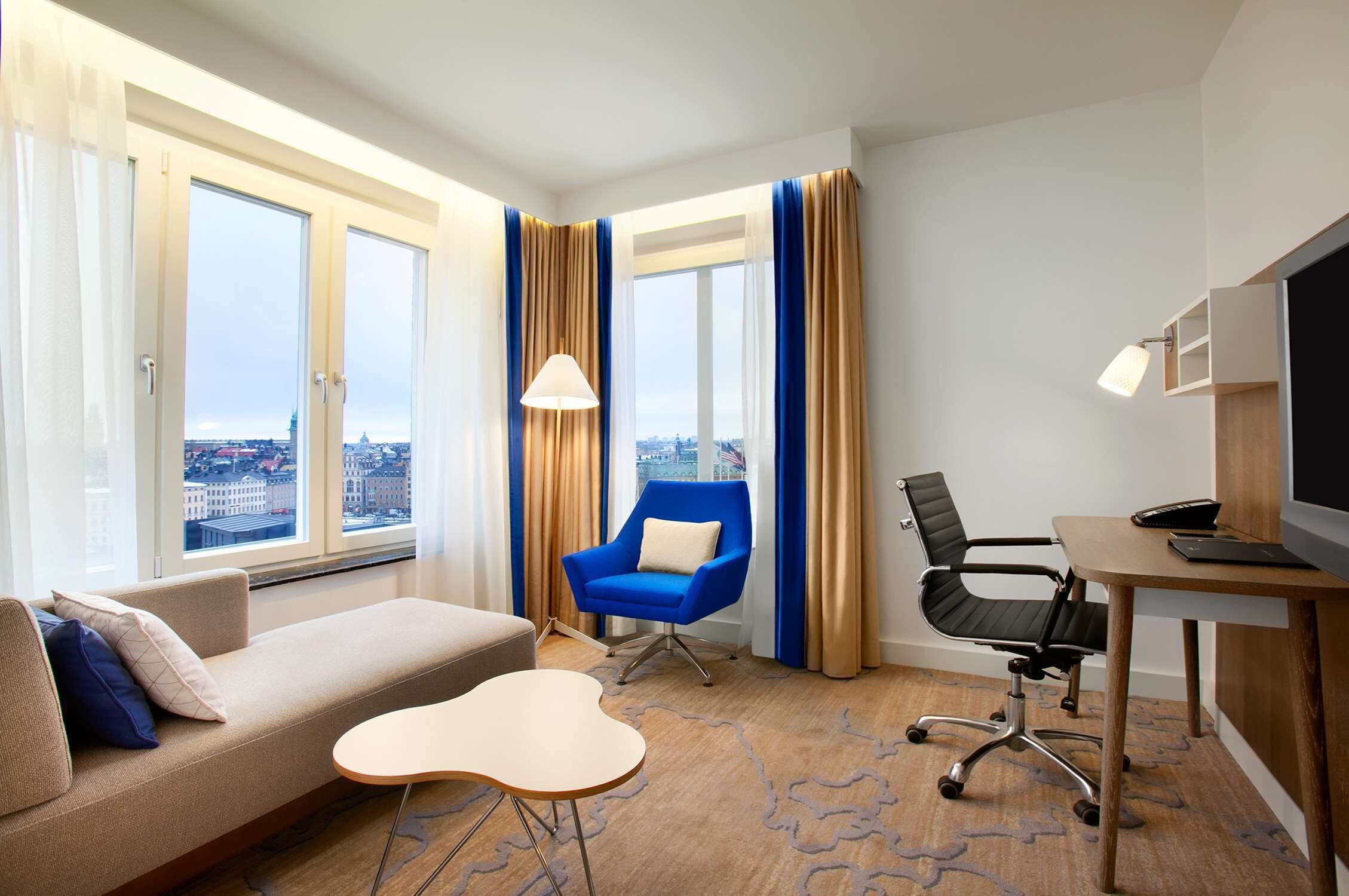 Una camera d'albergo con un divano, una poltrona blu, una scrivania con un computer e una televisione.