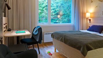Een hotelkamer met een bed, bureau, stoel, radiator en raam met uitzicht op groen.