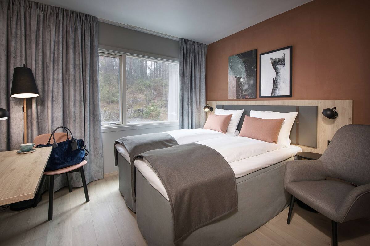 Habitación de hotel con una cama grande, un escritorio con una silla, una mesa con una taza y vistas al exterior a través de una ventana.