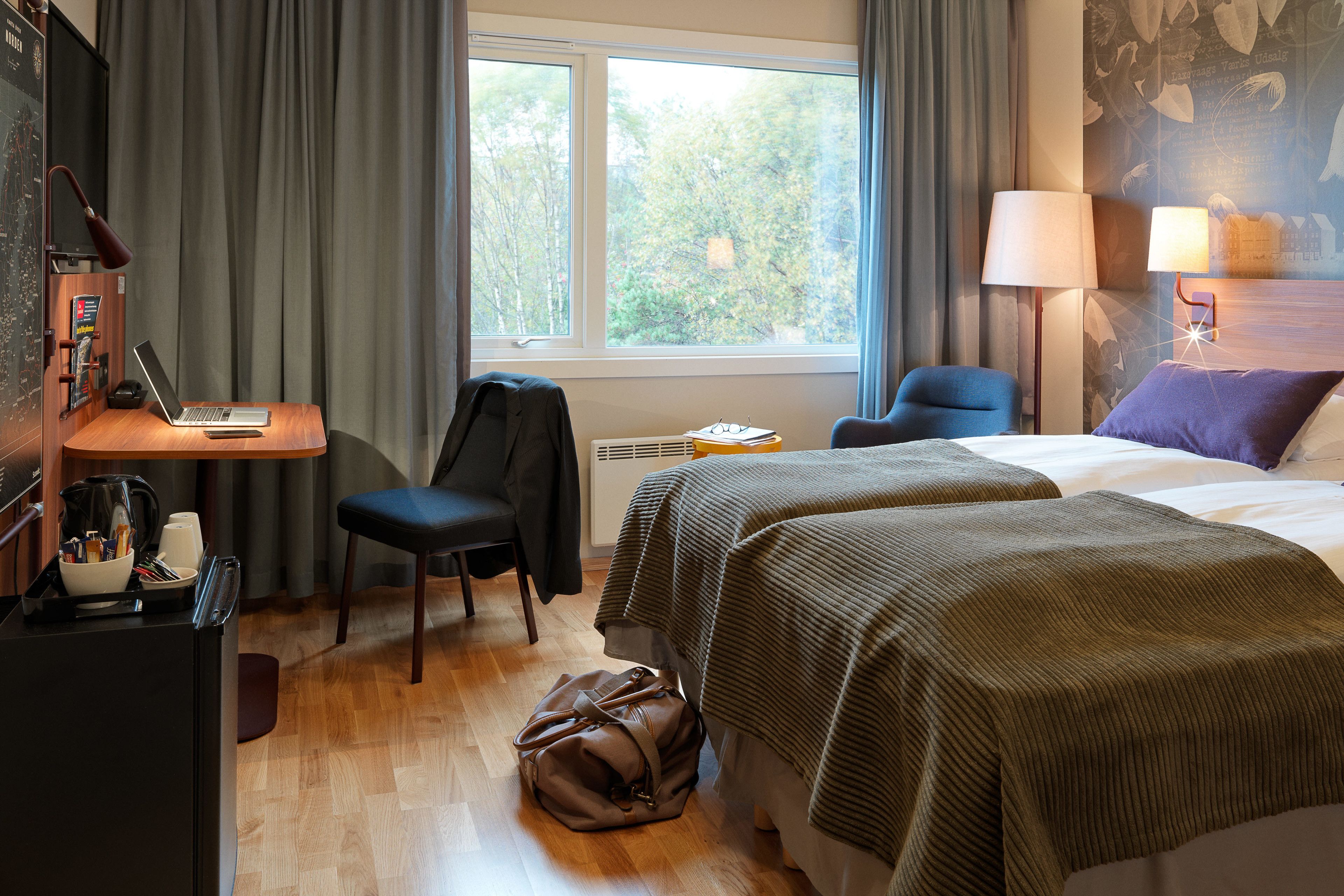 A hotel room with two beds, a desk with a laptop, a chair, a desk lamp, and a window with a view of trees.