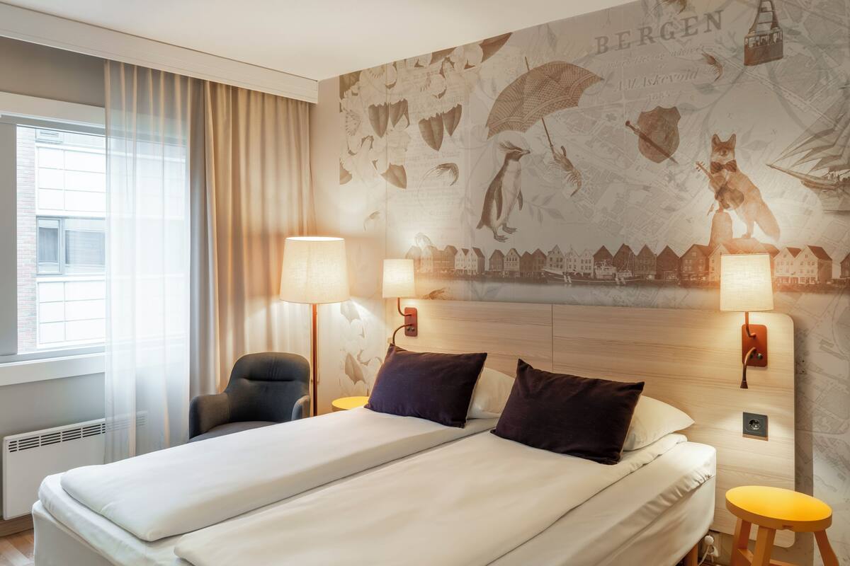 A hotel room with a bed, two bedside lamps, a chair, a small stool, and a wall with a map and illustrations.