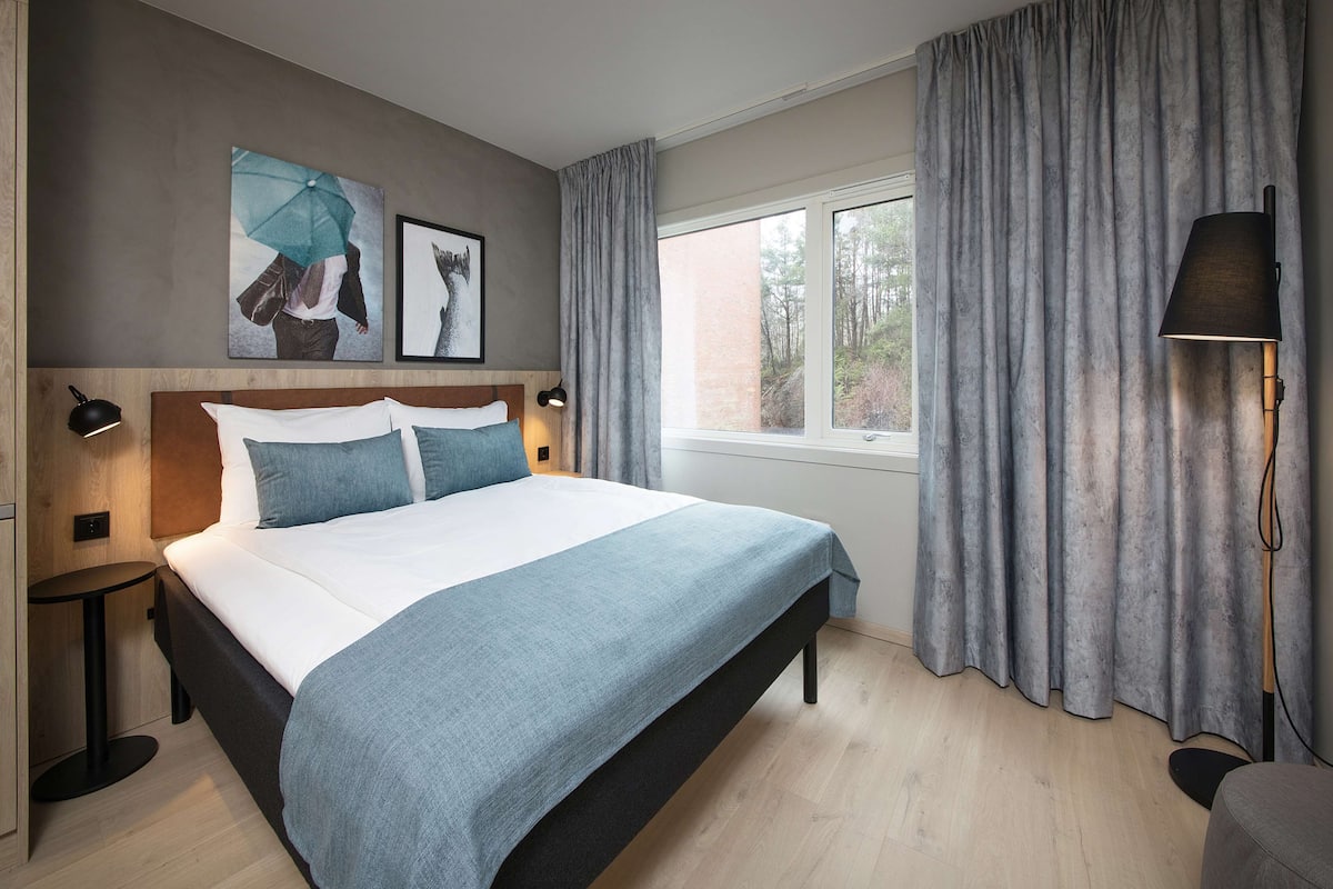 Un dormitorio moderno con una cama grande, una mesita de noche, una lámpara y una ventana con cortinas.