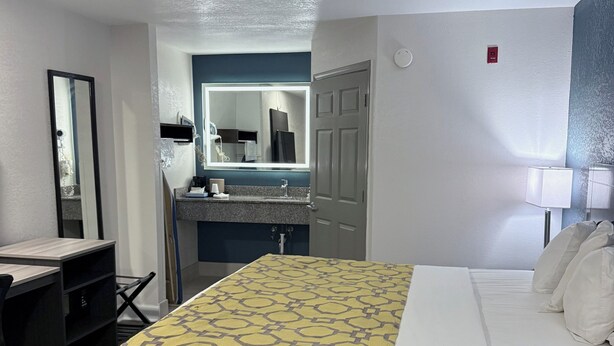 A hotel room with a bed, a desk, a mirror, a bathroom with a sink and mirror, and a door.