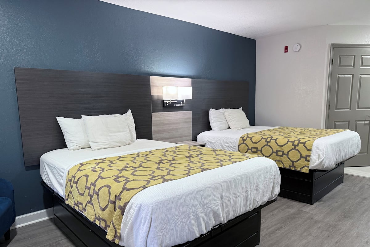 A hotel room with two beds, each with a white mattress and a patterned blanket. The room has a dark blue accent wall and a wooden headboard.