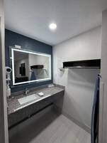 Un baño moderno con encimera de granito, un lavabo blanco, un espejo grande y una lámpara en el techo.
