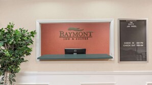 A reception desk with a sign for Baymont Inn & Suites, a plant, and a notice board with check-in and check-out times.