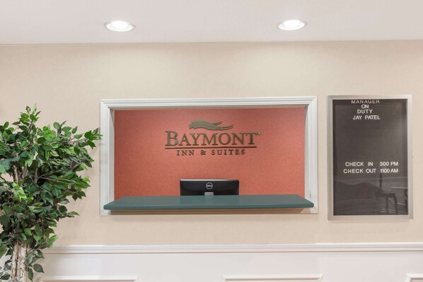 A reception desk with a sign for Baymont Inn & Suites, a plant, and a notice board with check-in and check-out times.