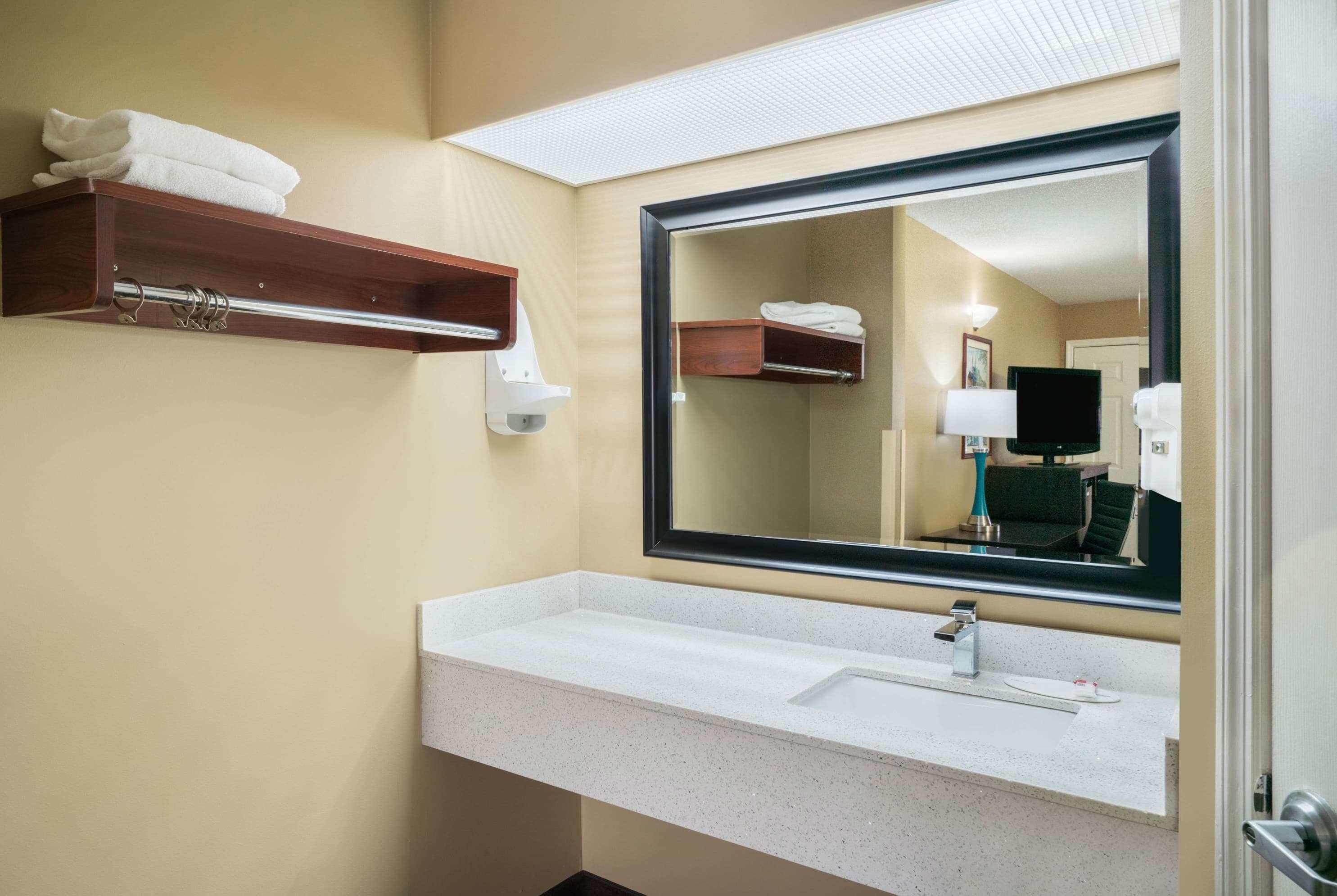 A hotel bathroom with a white sink, a large mirror, a towel rack, and a small wooden shelf.