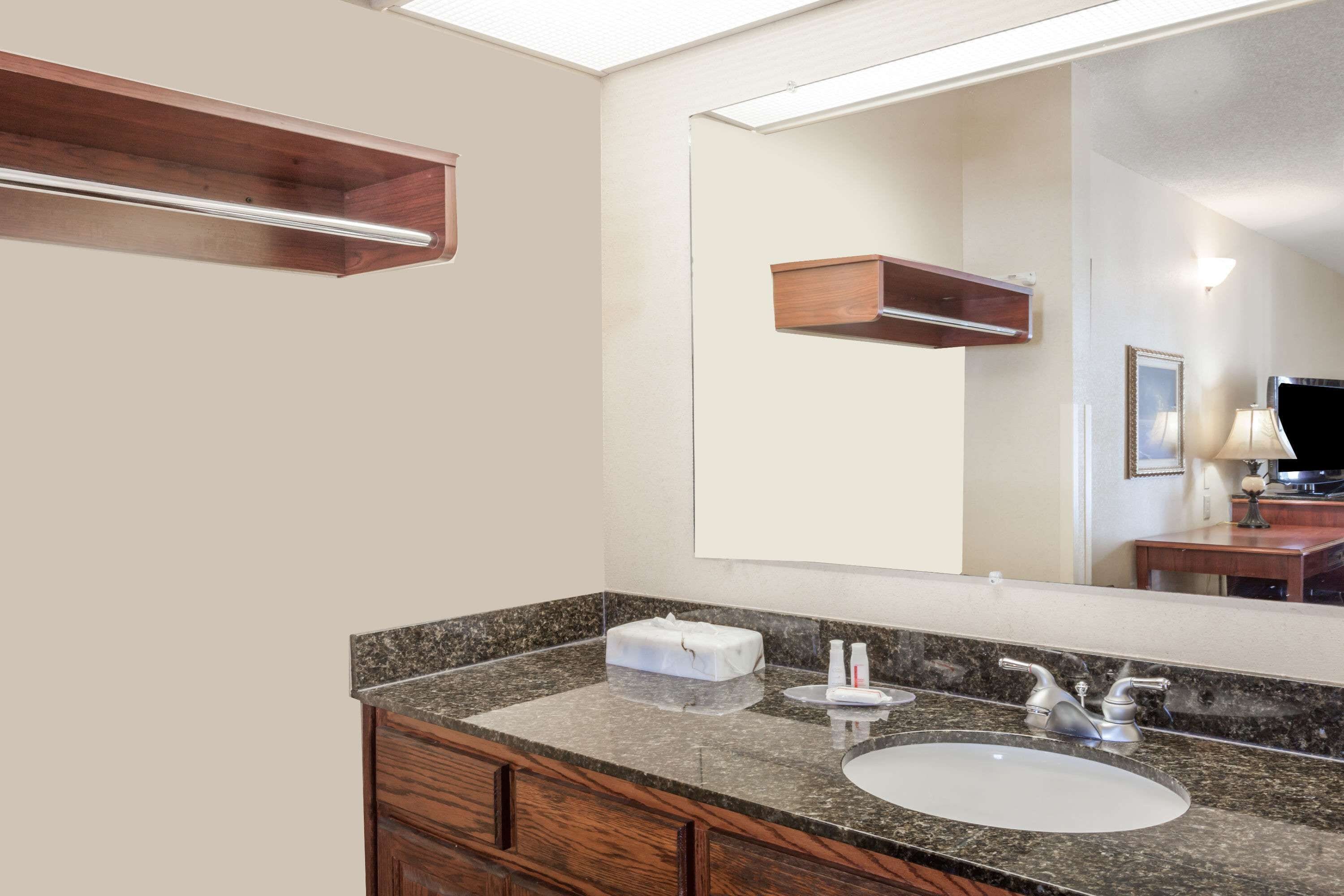 A bathroom with a granite countertop, a white sink, wooden cabinets, a mirror, and a wooden shelf above the mirror.