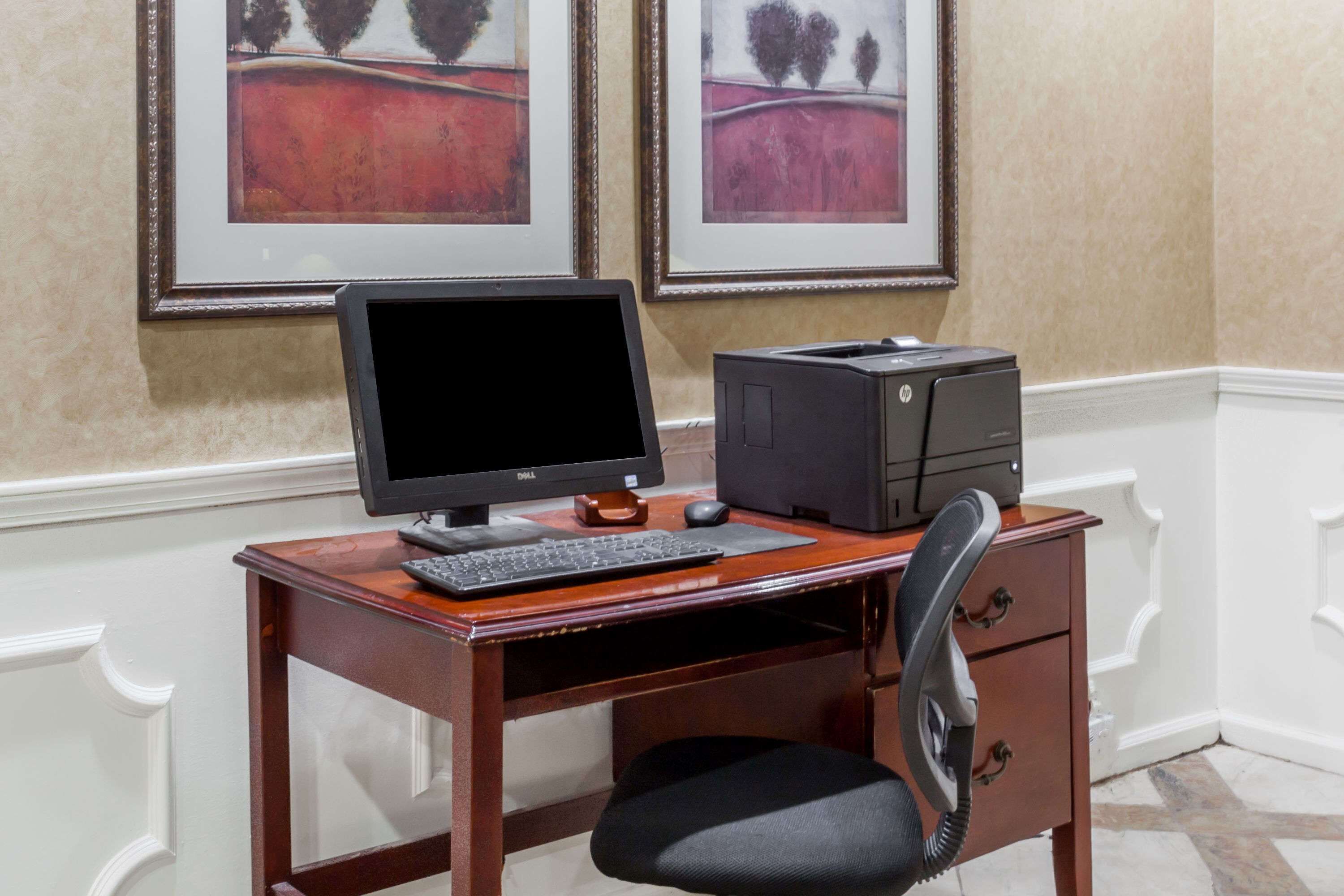 A wooden desk with a computer setup including a monitor, keyboard, mouse, and printer. There is a chair with a headrest. The desk is against a wall with two framed paintings above it.
