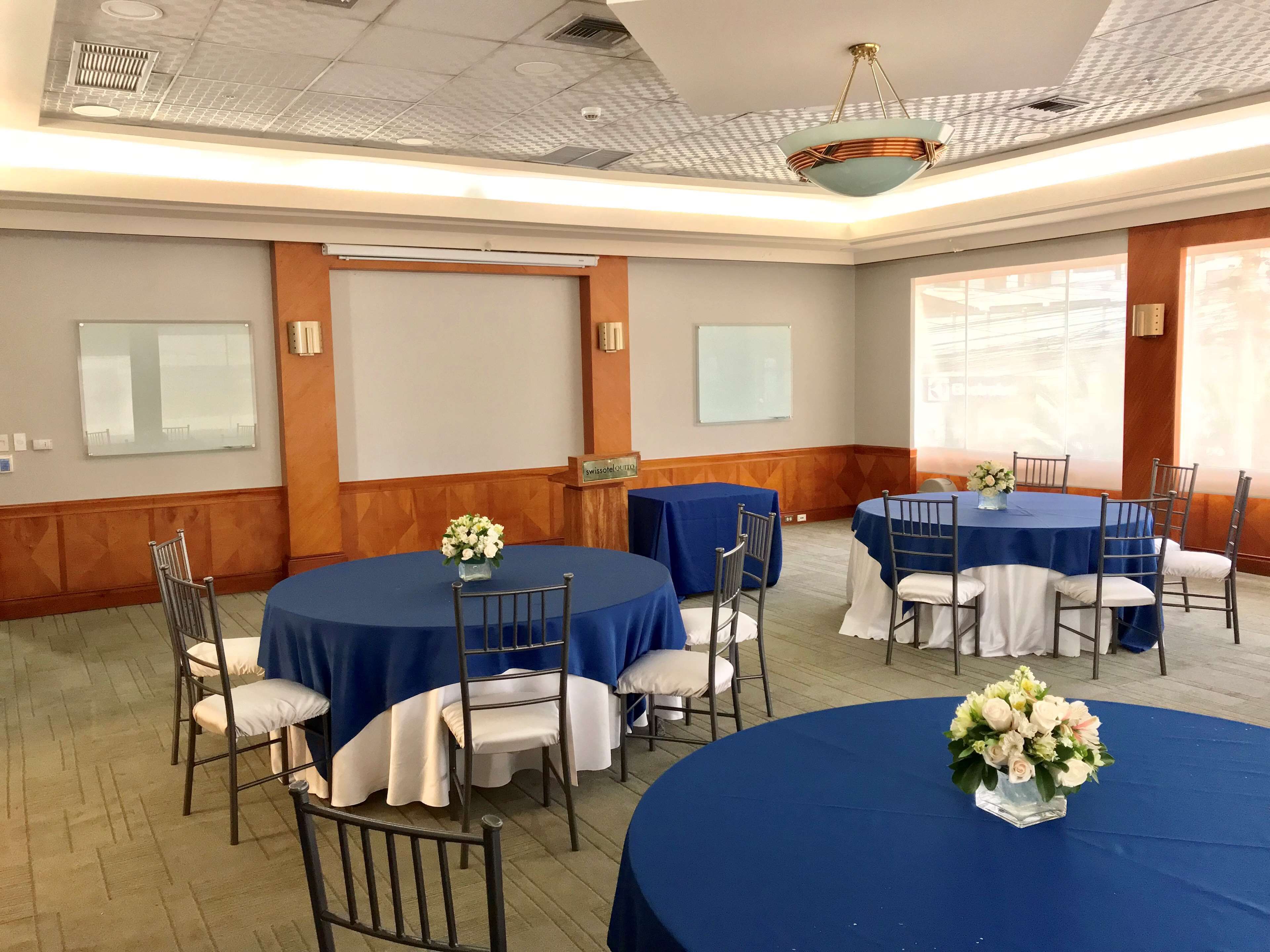 Un salón de conferencias con mesas redondas, sillas blancas y manteles azules. Hay arreglos florales blancos sobre las mesas.