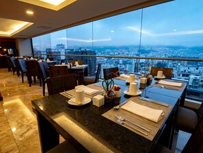 Un comedor con vista a la ciudad, mesas dispuestas con platos, cubiertos y servilletas.