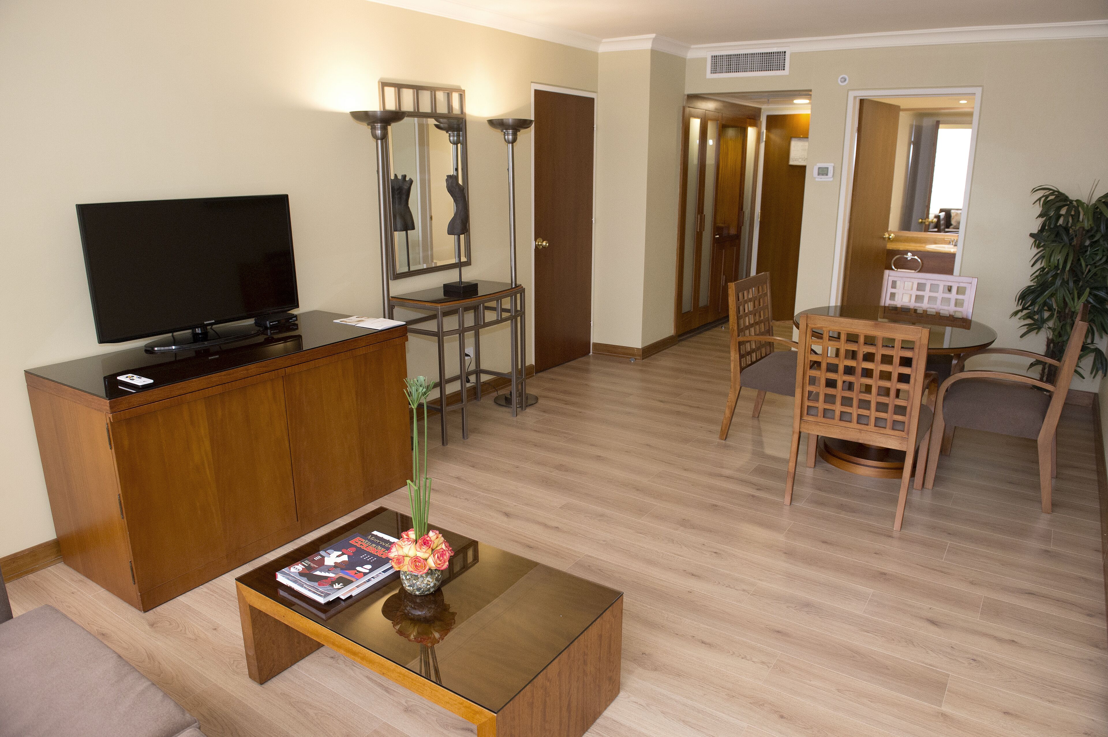 A living room with a flat-screen TV, wooden cabinet, coffee table with magazine and vase, and a dining area with chairs and a table.