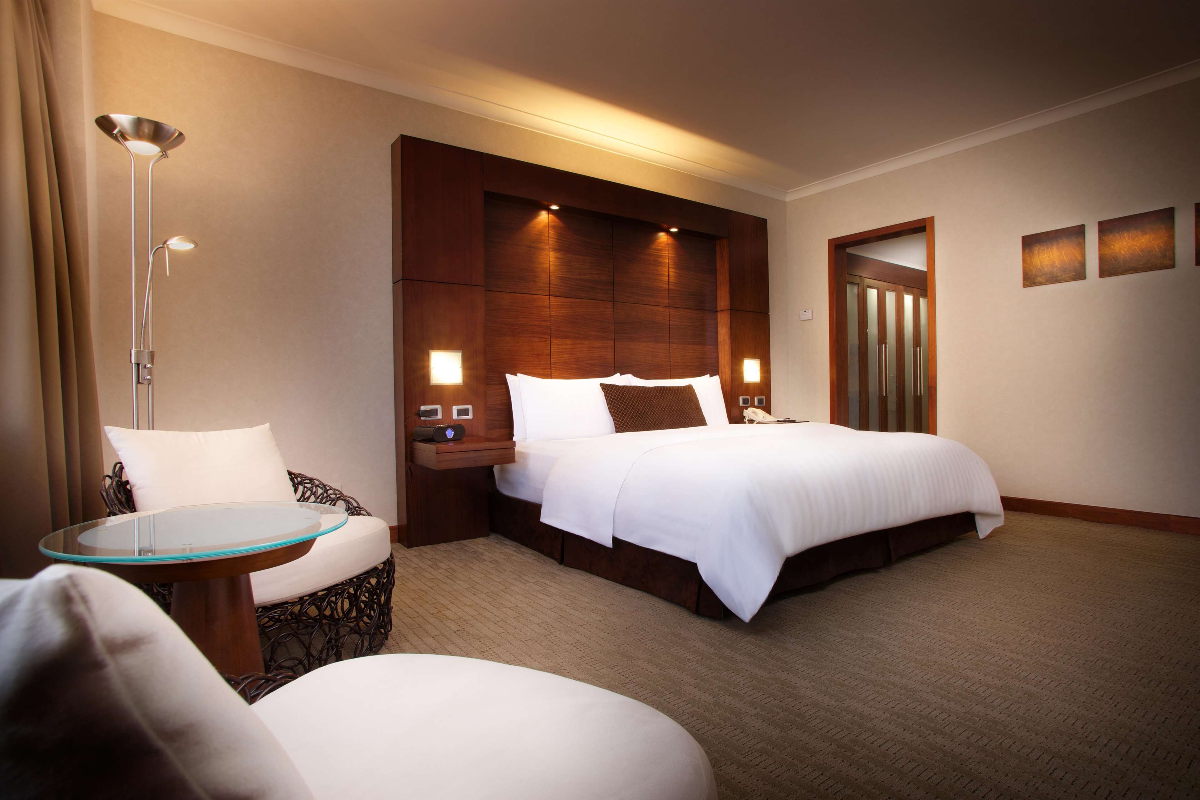 A hotel room with a large bed, a glass-top table, a chair, and a bedside table with a lamp.