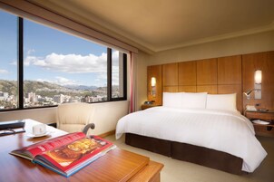 Une chambre d’hôtel avec un grand lit, un bureau avec un magazine, une chaise et une vue sur le paysage urbain à travers de grandes fenêtres.