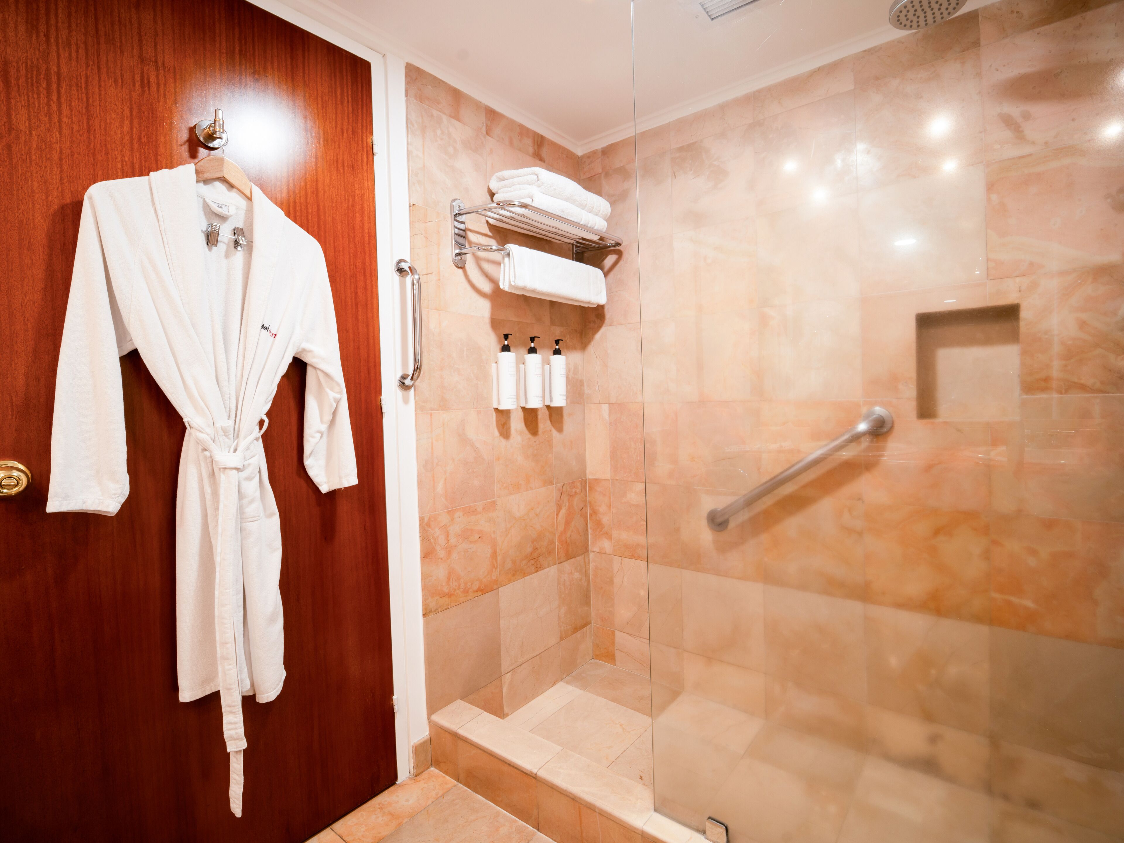 A bathroom with a wooden door, a white robe hanging on a rack, a glass shower enclosure, and a towel rack with folded towels.