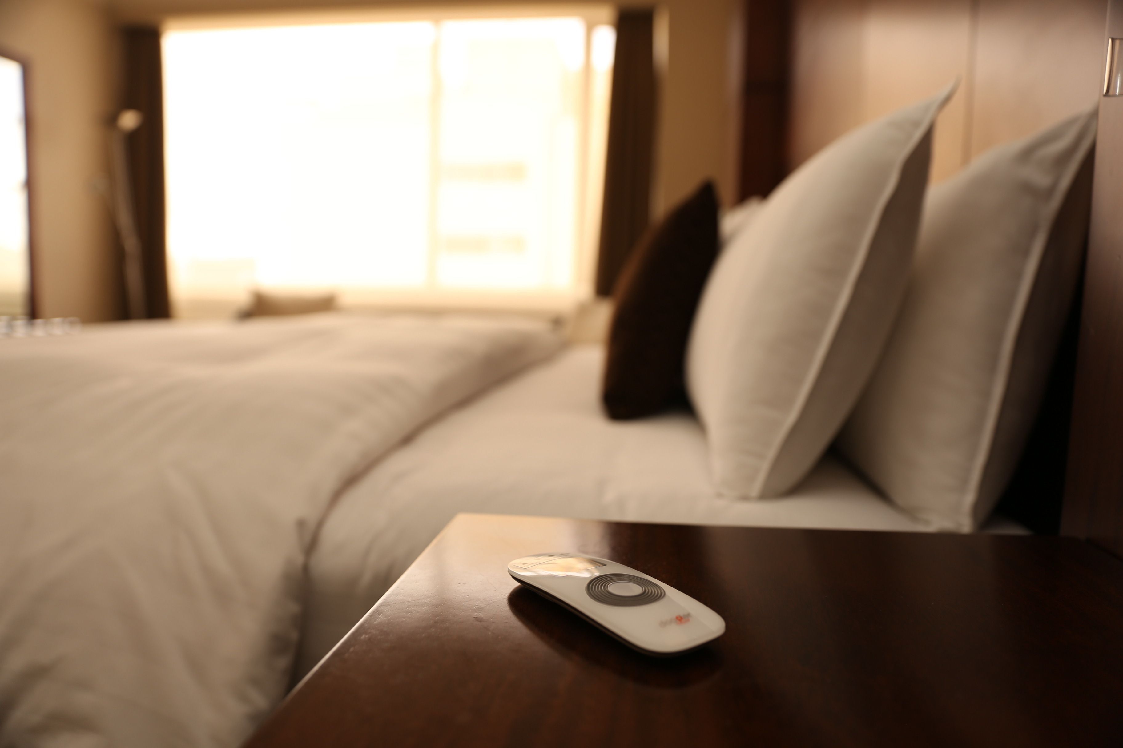 A hotel room with a bed, pillows, and a remote control on a wooden nightstand.