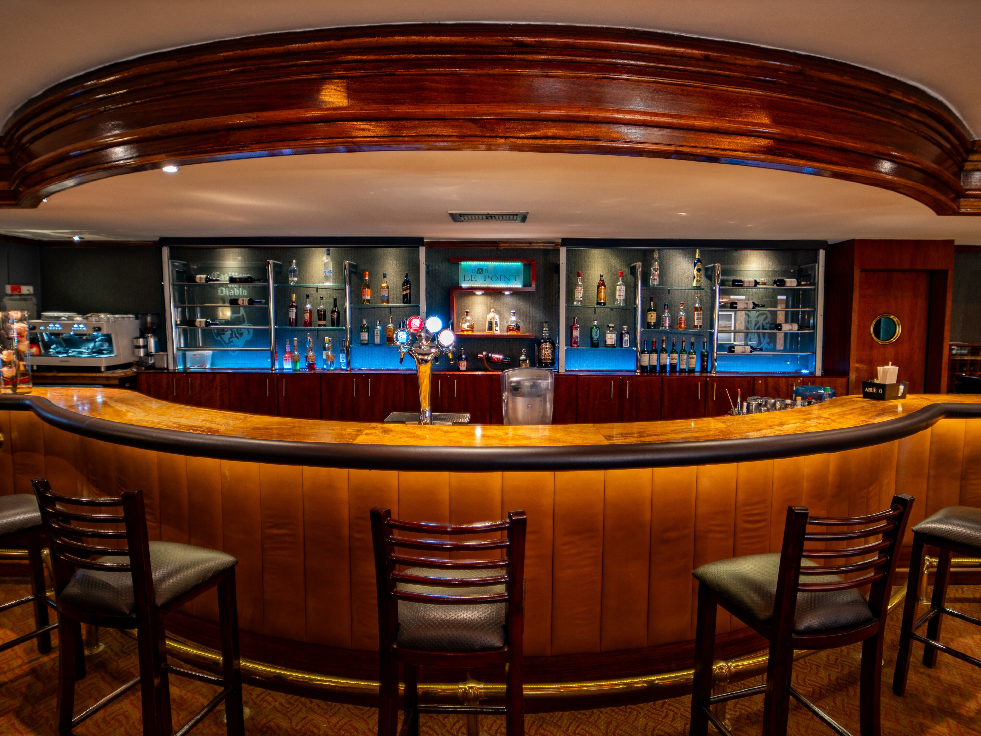A bar area with a curved wooden counter, multiple shelves of bottles, and three bar stools.