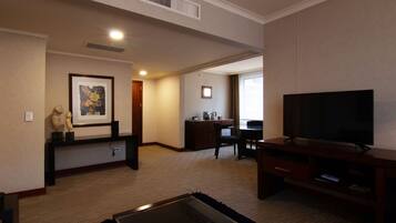 A hotel room with a flat-screen TV, a small coffee table, a framed artwork, a desk with a chair, and a kitchenette area.