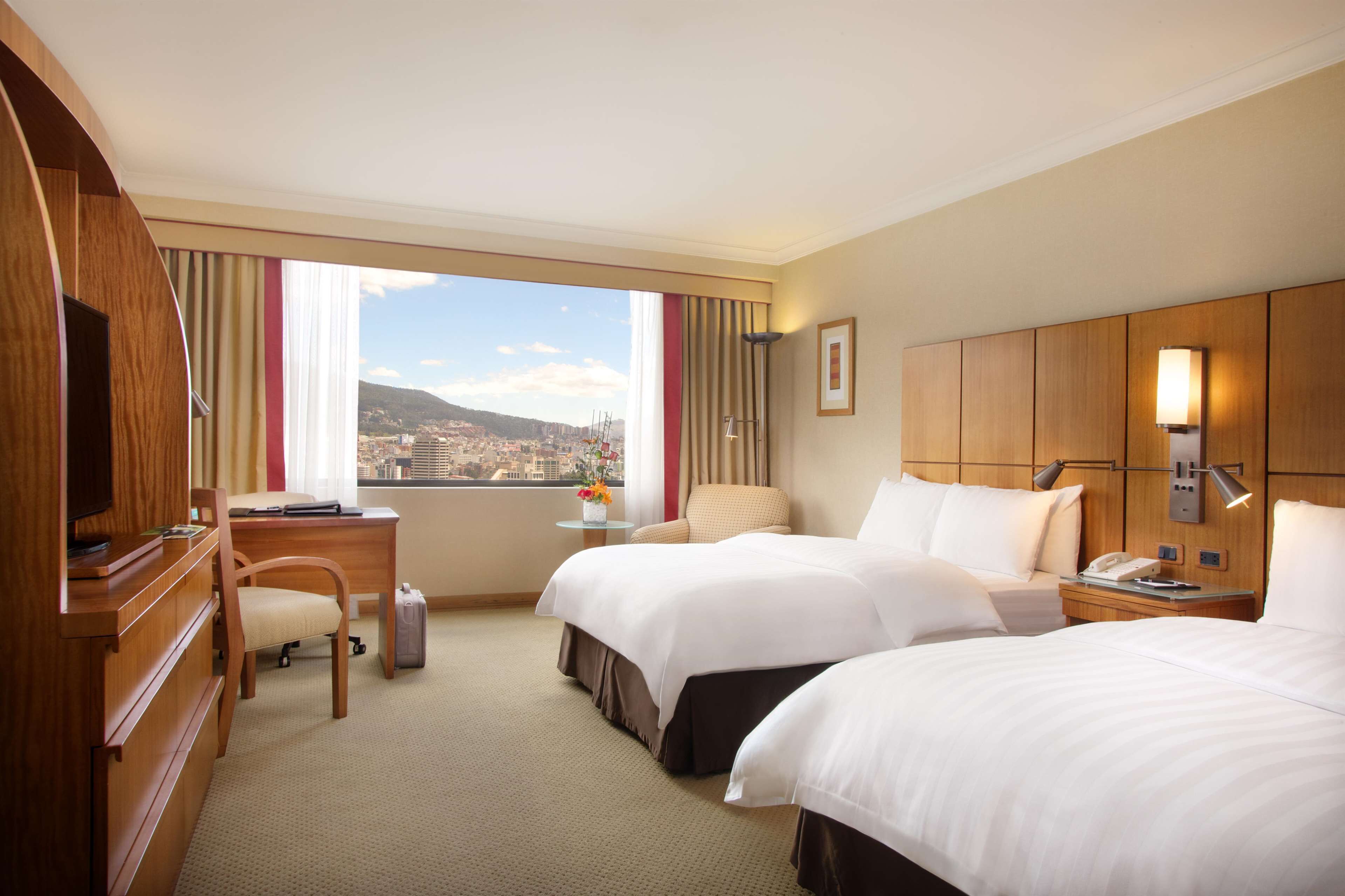 A hotel room with two beds, a desk, a chair, a TV, a window with a city view, and a bedside table with a lamp.