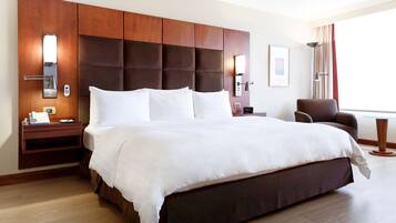 A hotel room with a large bed, wooden headboard, bedside tables with lamps, a chair, and a large window with curtains.