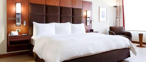 A hotel room with a large bed, wooden headboard, bedside tables with lamps, a chair, and a large window with curtains.