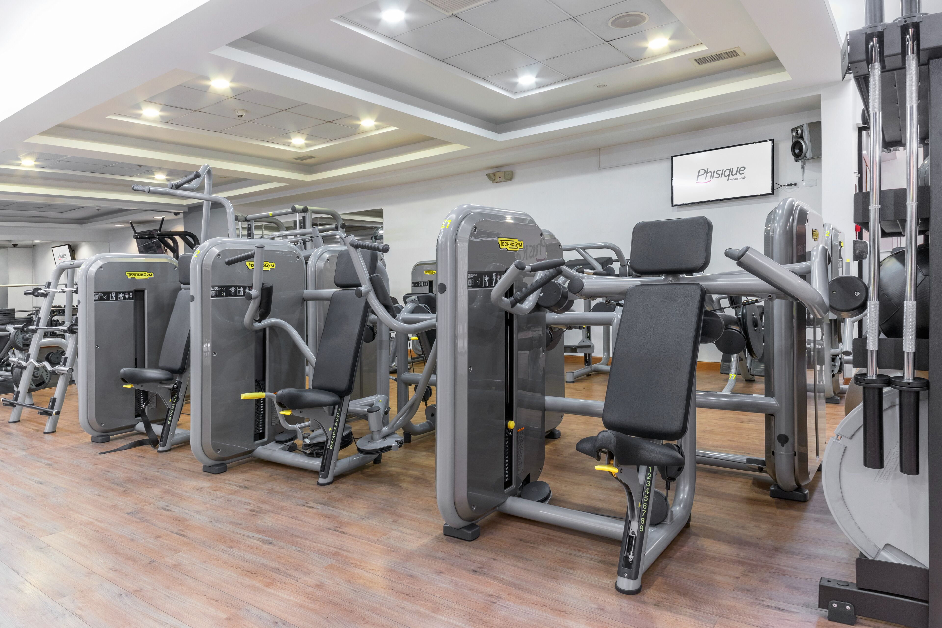Un gimnasio con diversas máquinas de ejercicio, un techo blanco con iluminación empotrada y un letrero que dice 'Physique'.