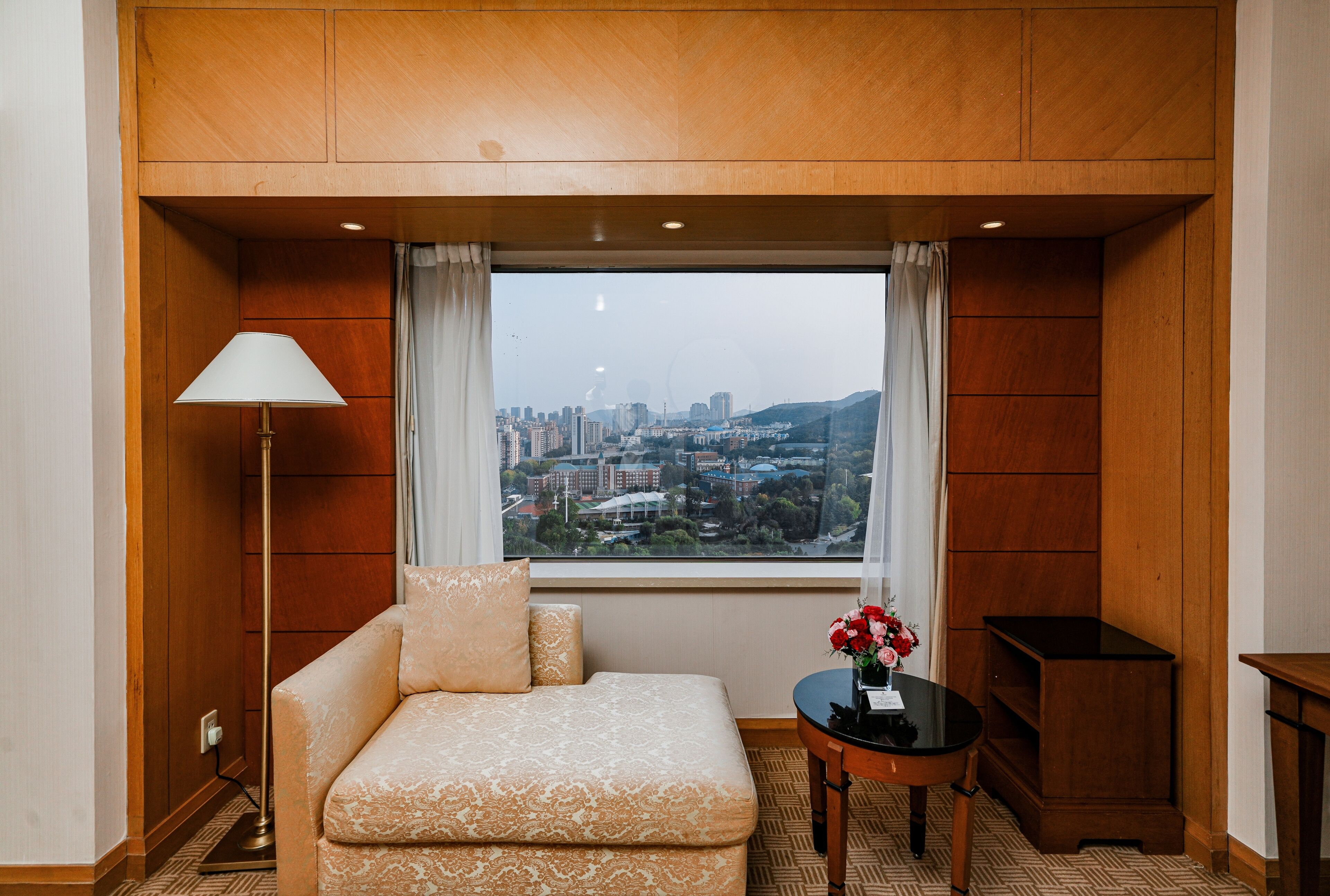 A hotel room with a sofa, a small table with a vase, and a view of a cityscape through large windows.