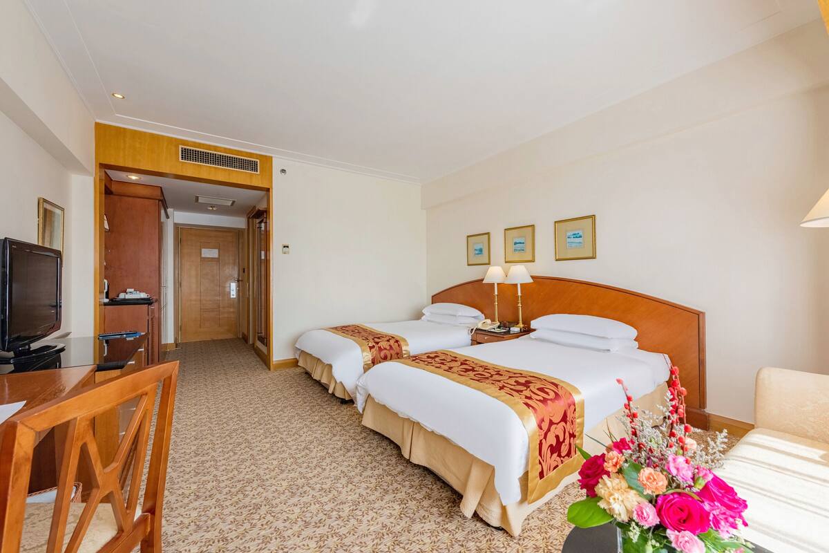 A hotel room with two beds, a desk with a TV, and a chair. There are framed pictures on the wall and a vase of flowers on the bed.