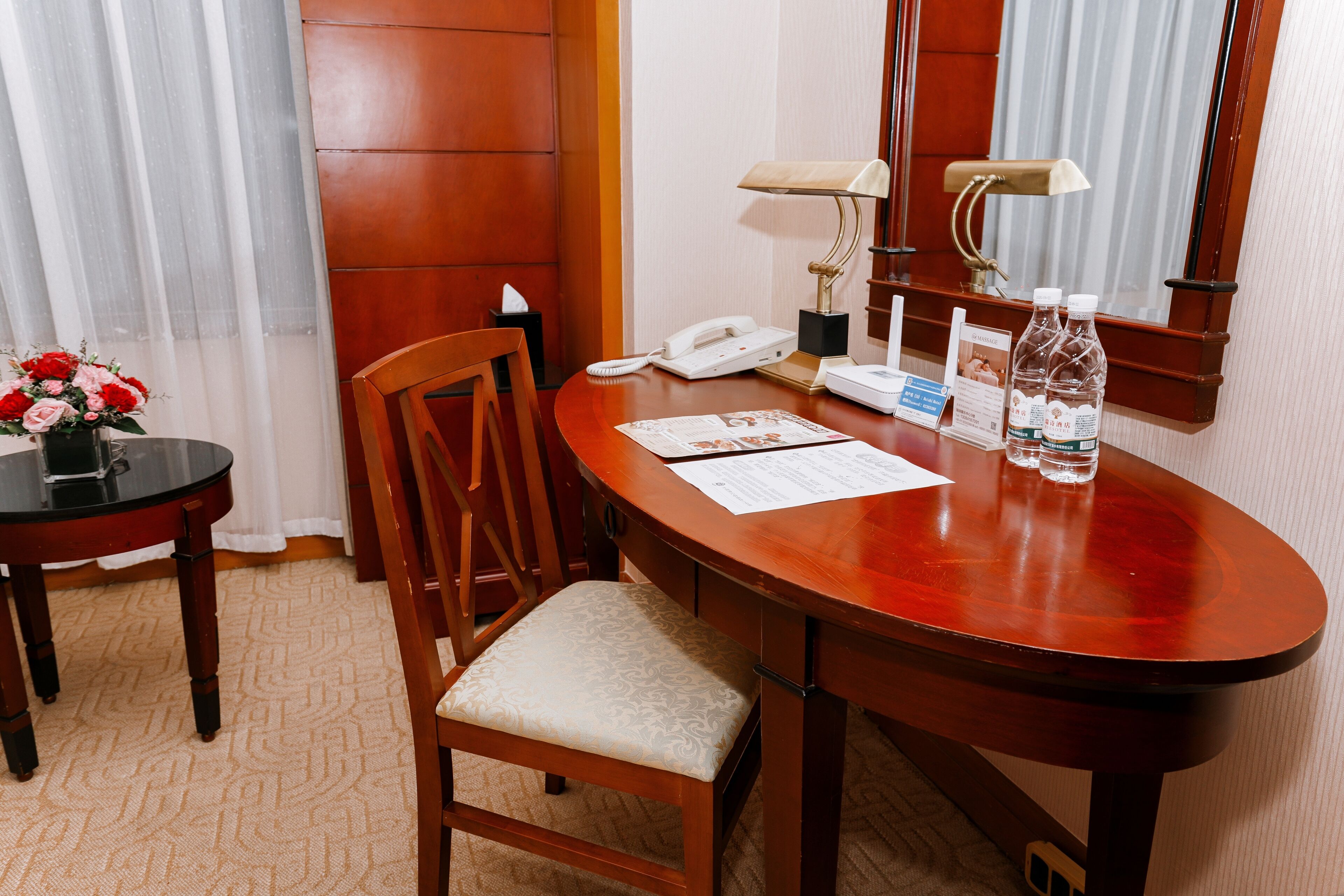 A hotel room with a wooden desk, a telephone, a lamp, a chair, and a small table with a vase of flowers.
