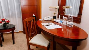 A hotel room with a wooden desk, a telephone, a lamp, a chair, and a small table with a vase of flowers.