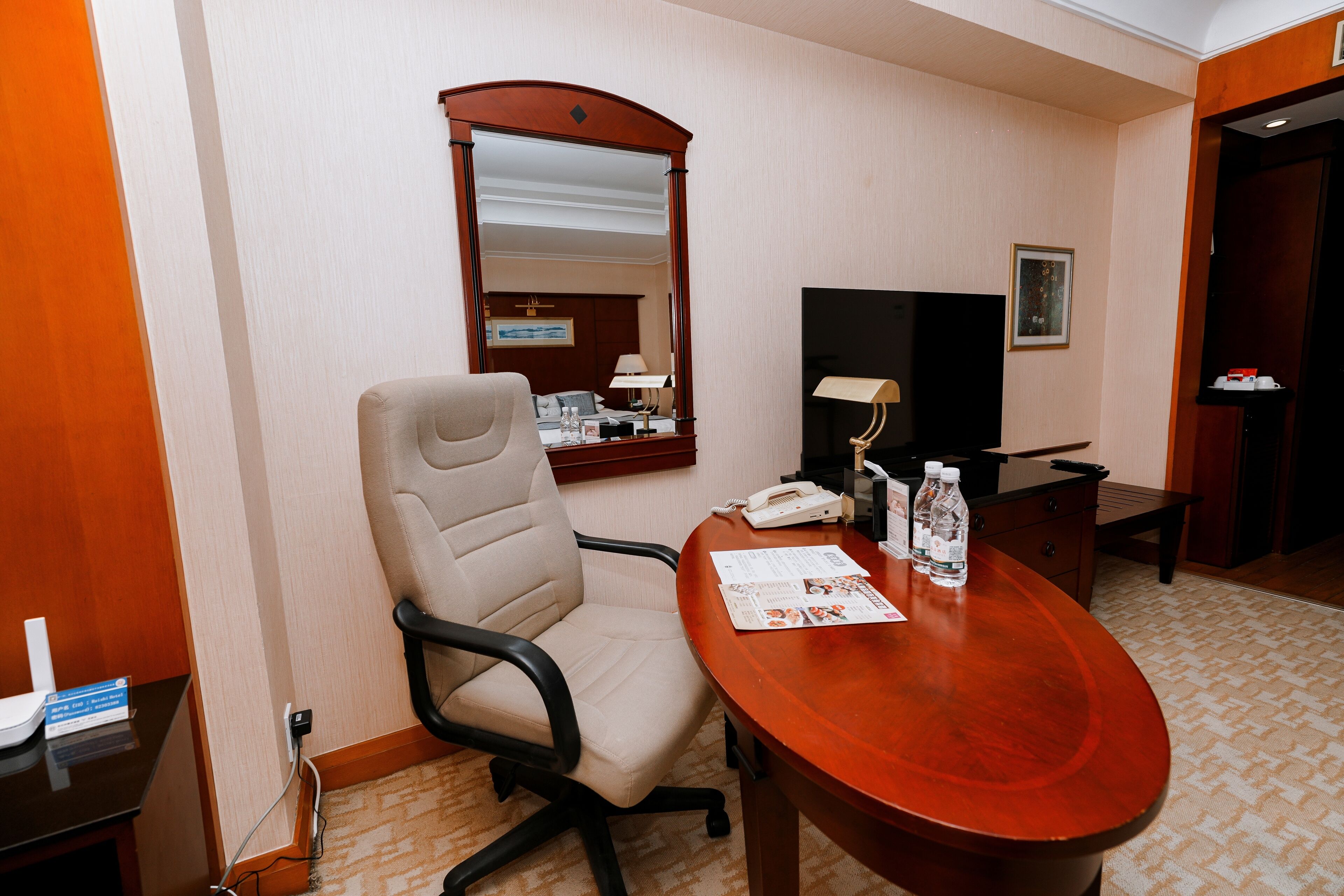 Une chambre d’hôtel avec un bureau en bois, une chaise, un miroir, une télévision et une petite table avec une lampe et une bouteille d’eau.
