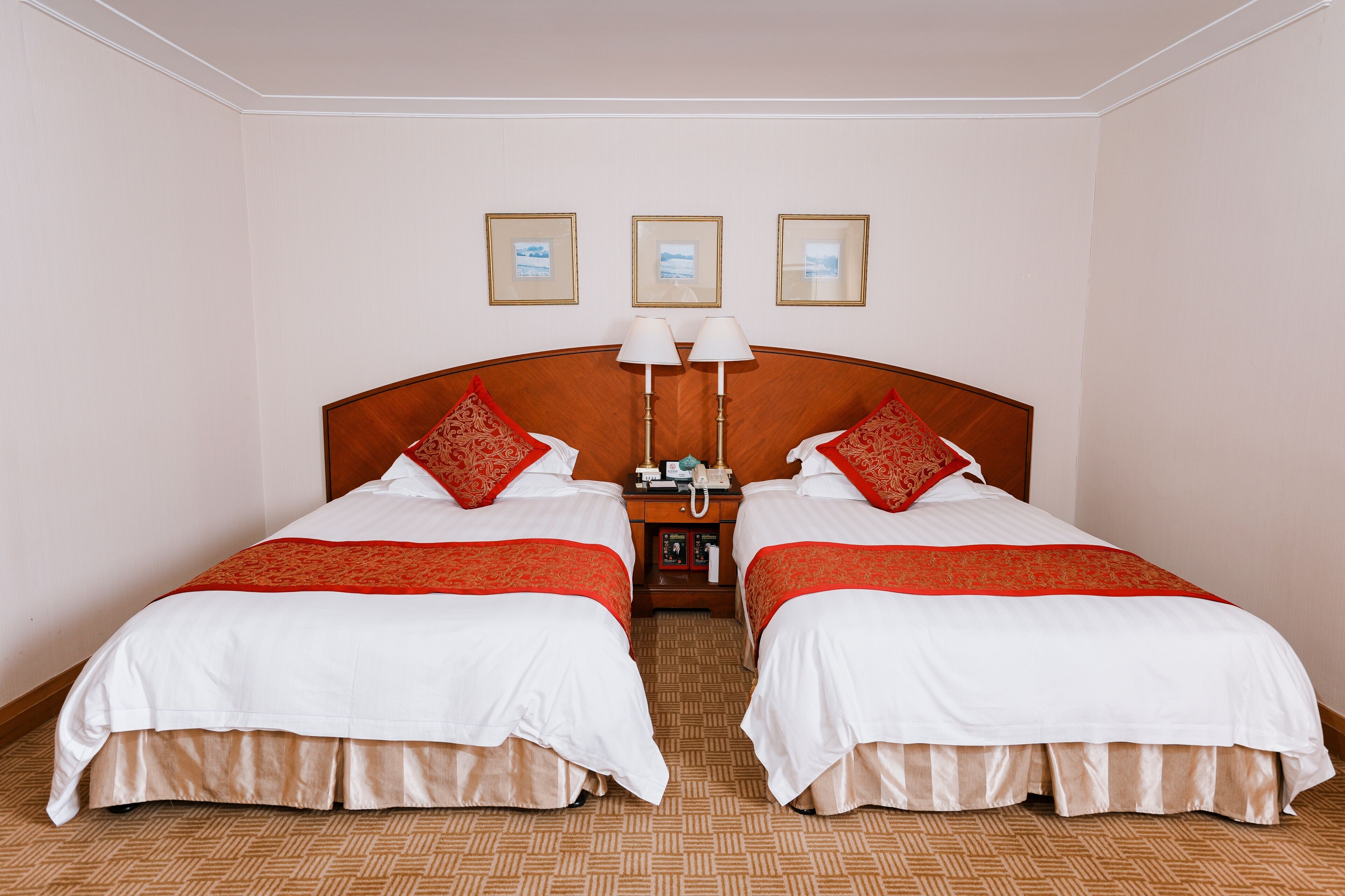Habitación de hotel con dos camas, cada una con ropa de cama roja y blanca, cabeceros de madera y mesitas de noche con lámparas.