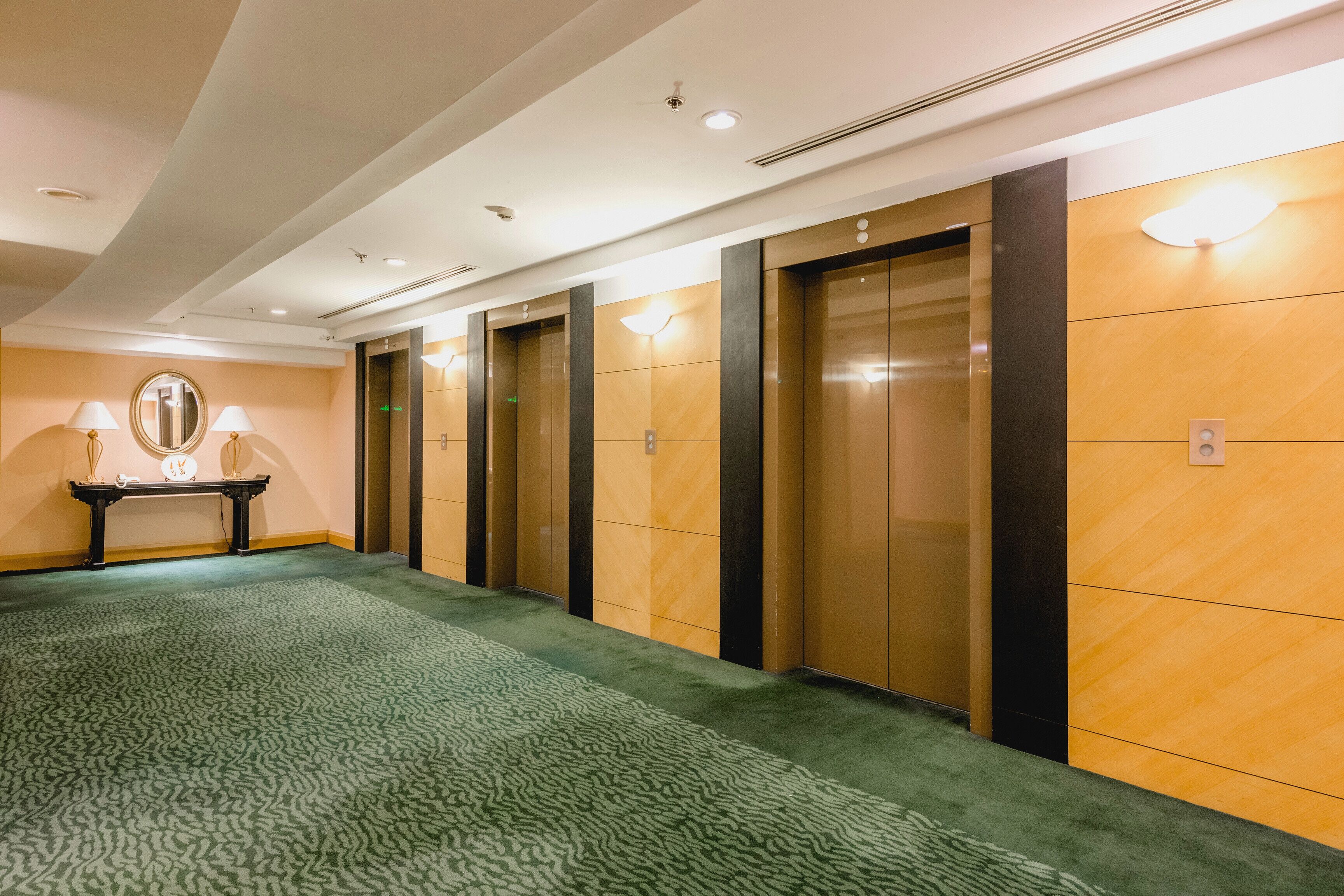 A hotel lobby with a green carpet, a round mirror, and three elevators.