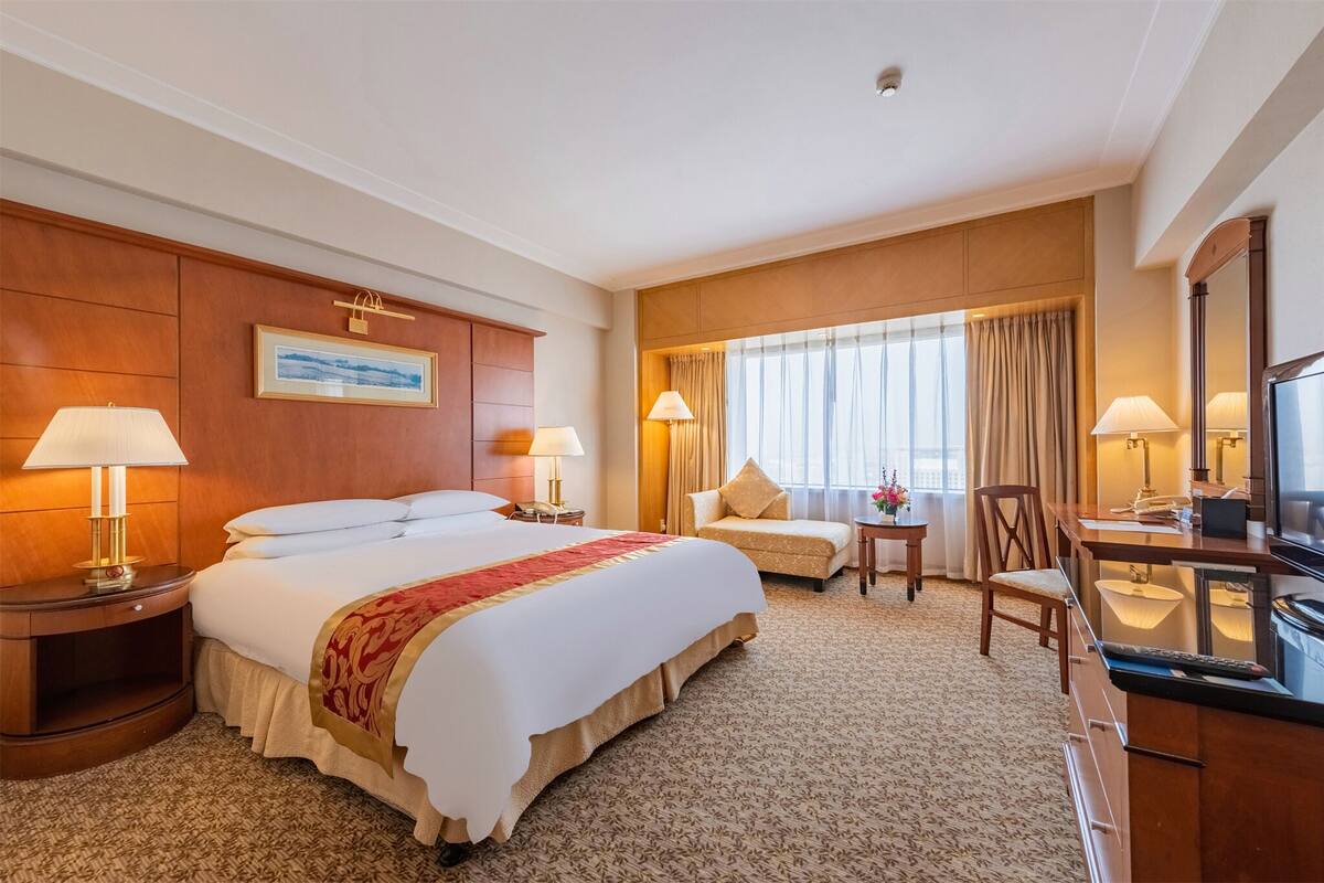 A hotel room with a large bed, two bedside tables with lamps, a small table with a chair, a desk with a chair, a large window with curtains, and a framed picture on the wall.