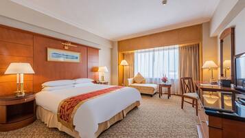 Habitación de hotel con una cama grande, dos mesitas de noche con lámparas, una pequeña mesa con una silla, un escritorio con silla, un ventanal con cortinas y un cuadro en la pared.