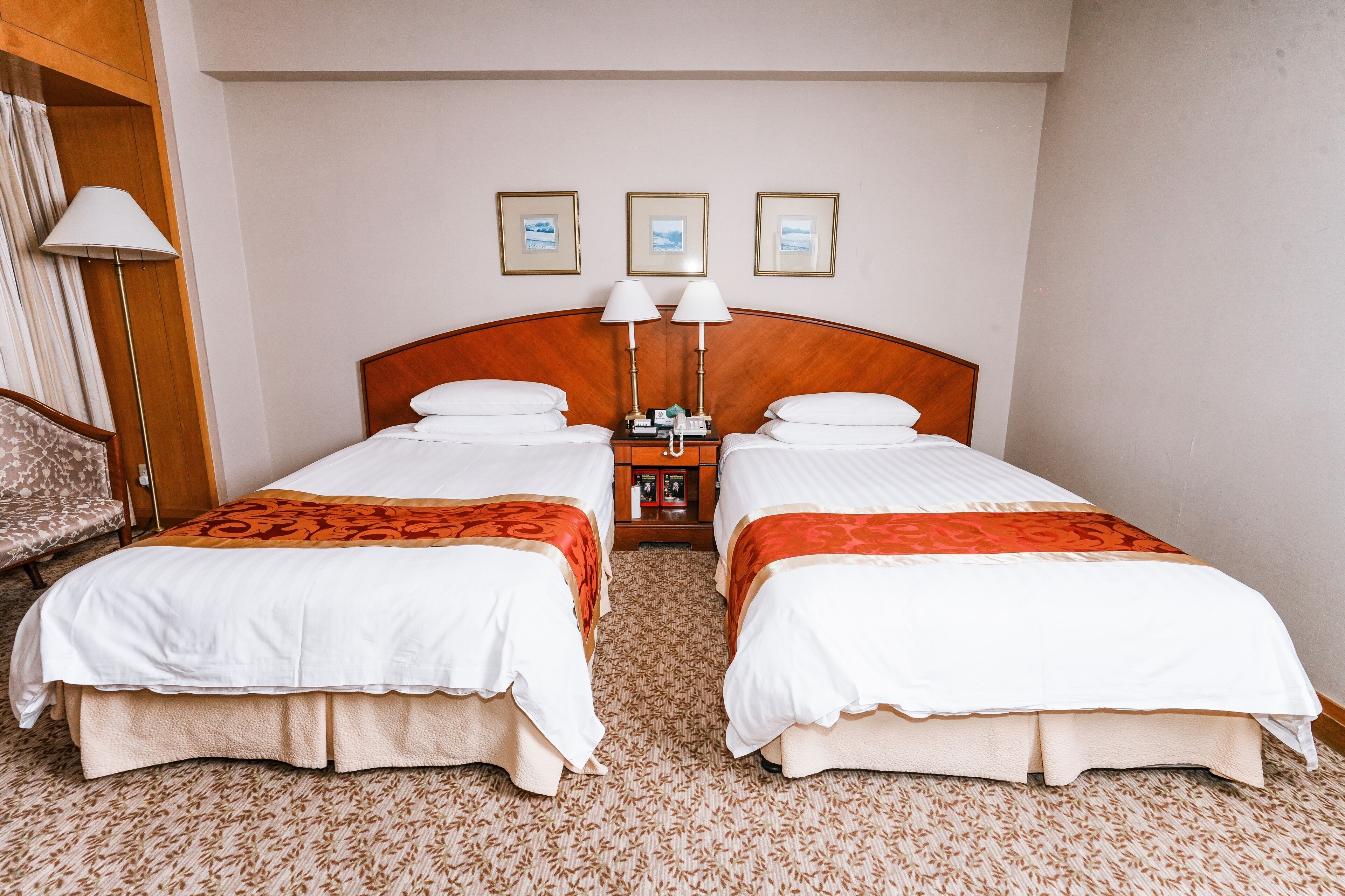 A hotel room with two beds, a nightstand with lamps, and three framed pictures on the wall.
