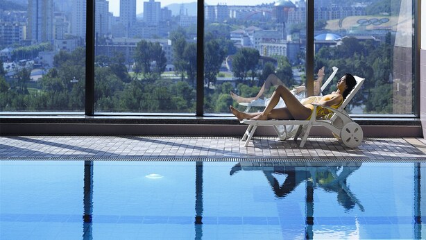 A person relaxing by a pool with a cityscape view through large windows. - Ruishi Hotel Dalian (Dalian)