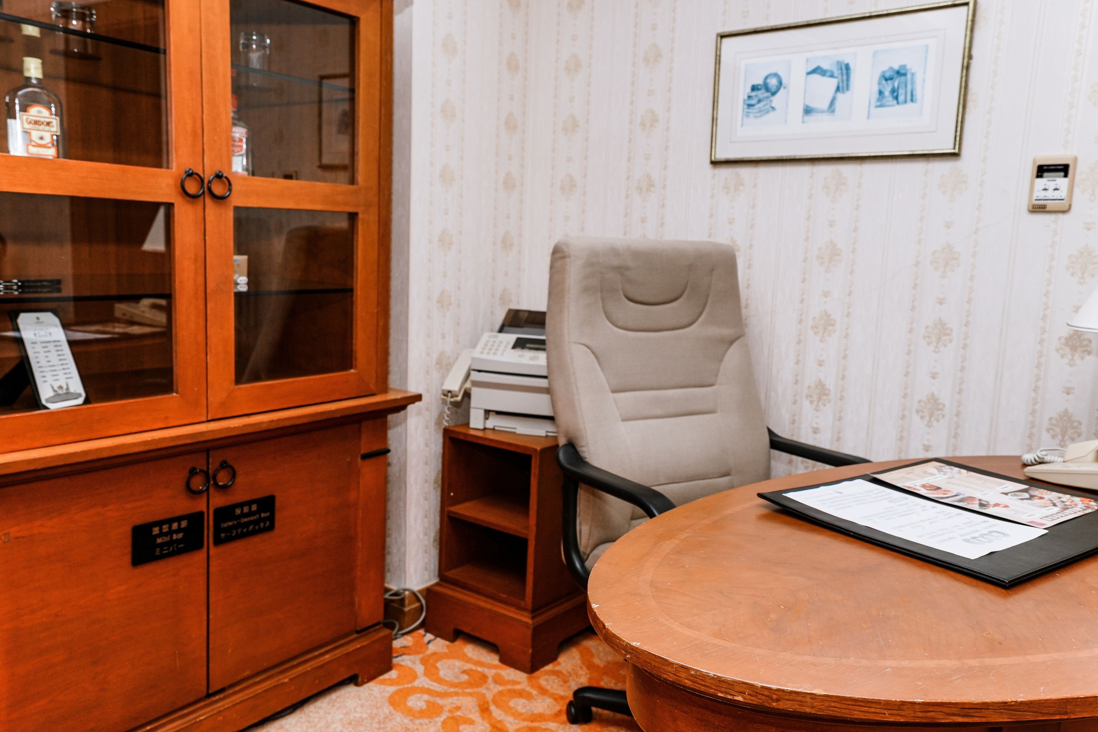 A room with a wooden cabinet, a beige office chair, a round wooden table with a menu, and a patterned wallpaper.