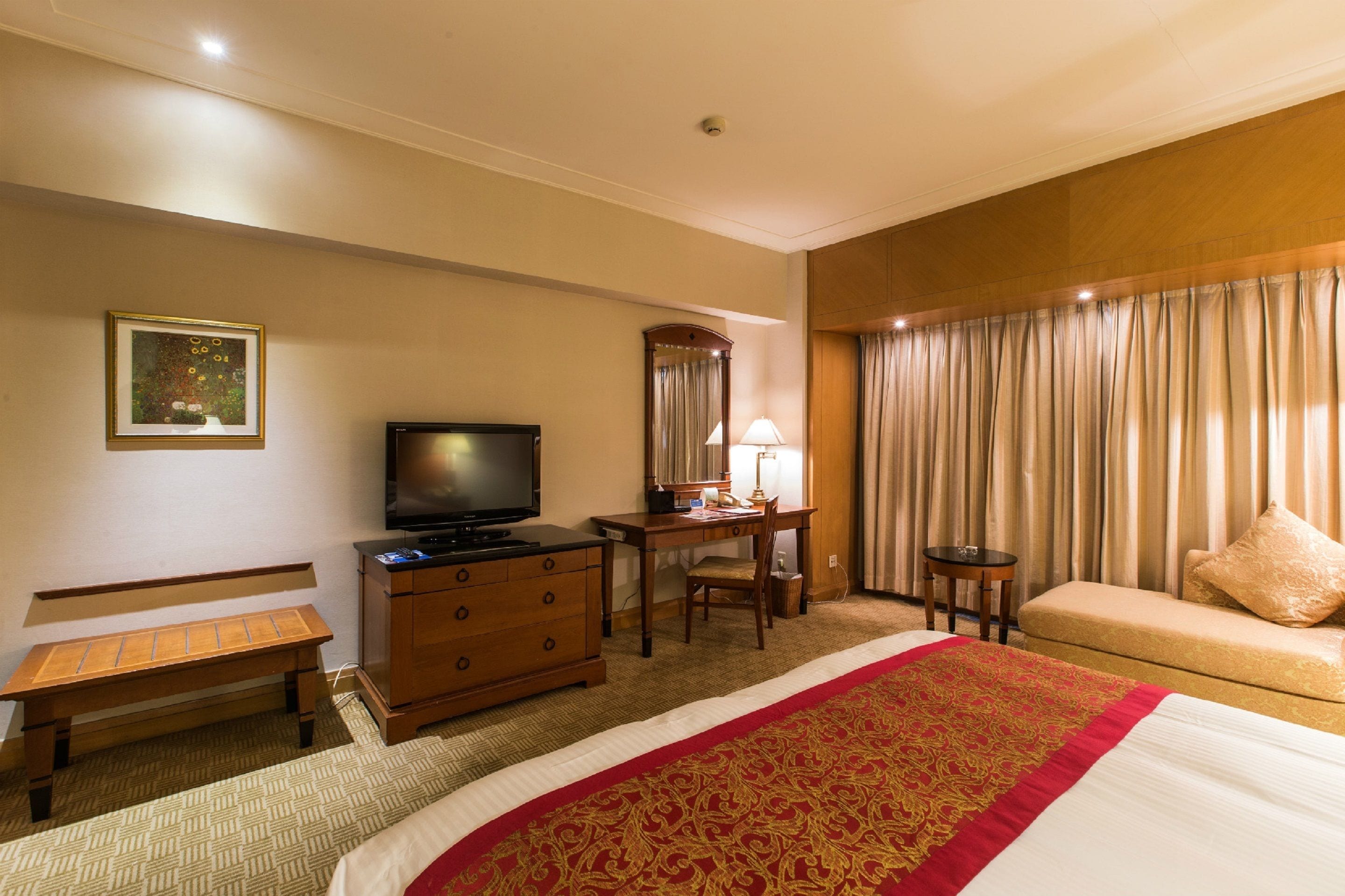 A hotel room with a bed, a desk with a computer, a television, a bench, a dresser, and a window with curtains.