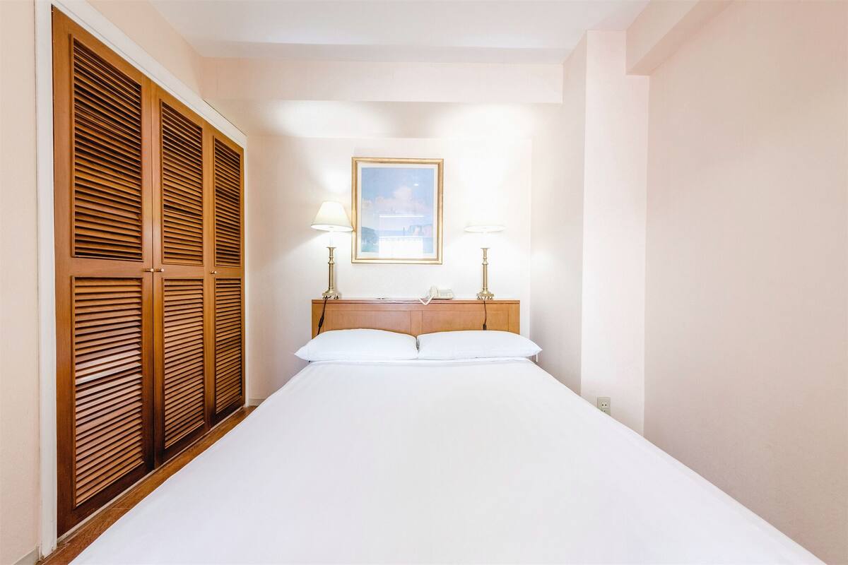 A hotel room with a bed, wooden wardrobe, two bedside lamps, and a framed picture on the wall.