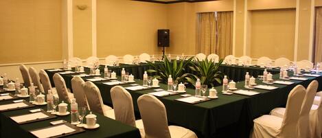 Una sala de conferencias con varias mesas, sillas y un orador. Las mesas están dispuestas con botellas de agua, vasos y blocs de notas.