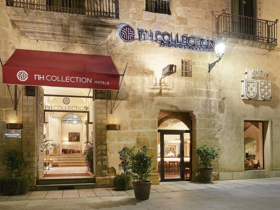 Entrada de un hotel con toldo rojo, un cartel que dice 'NH COLLECTION Hotels' y fachada de piedra.