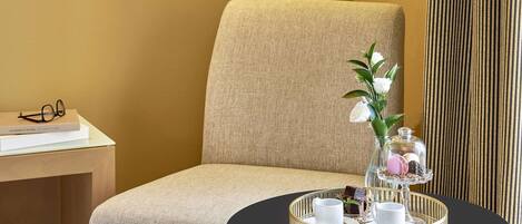 Habitación de hotel con un sillón tapizado en beige, una mesita redonda con una bandeja de aperitivos, un florero con flores blancas y un libro sobre una mesita auxiliar.