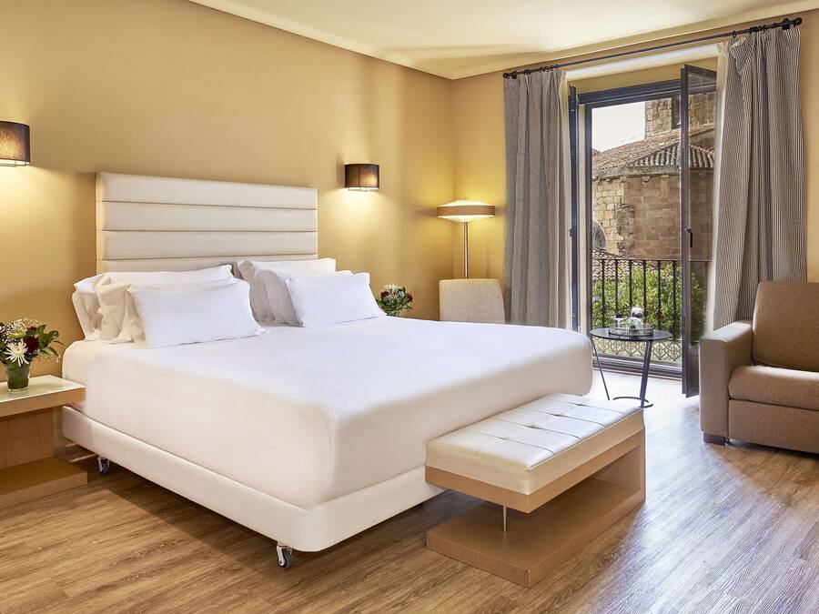 Habitación de hotel con una cama grande, mesitas de noche, un banco, un sofá y un balcón con vistas a un edificio.