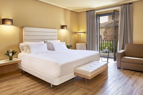 A hotel room with a large bed, bedside tables, a bench, a sofa, and a balcony with a view of a building. - NH Collection Cáceres Palacio de Oquendo (Cáceres)
