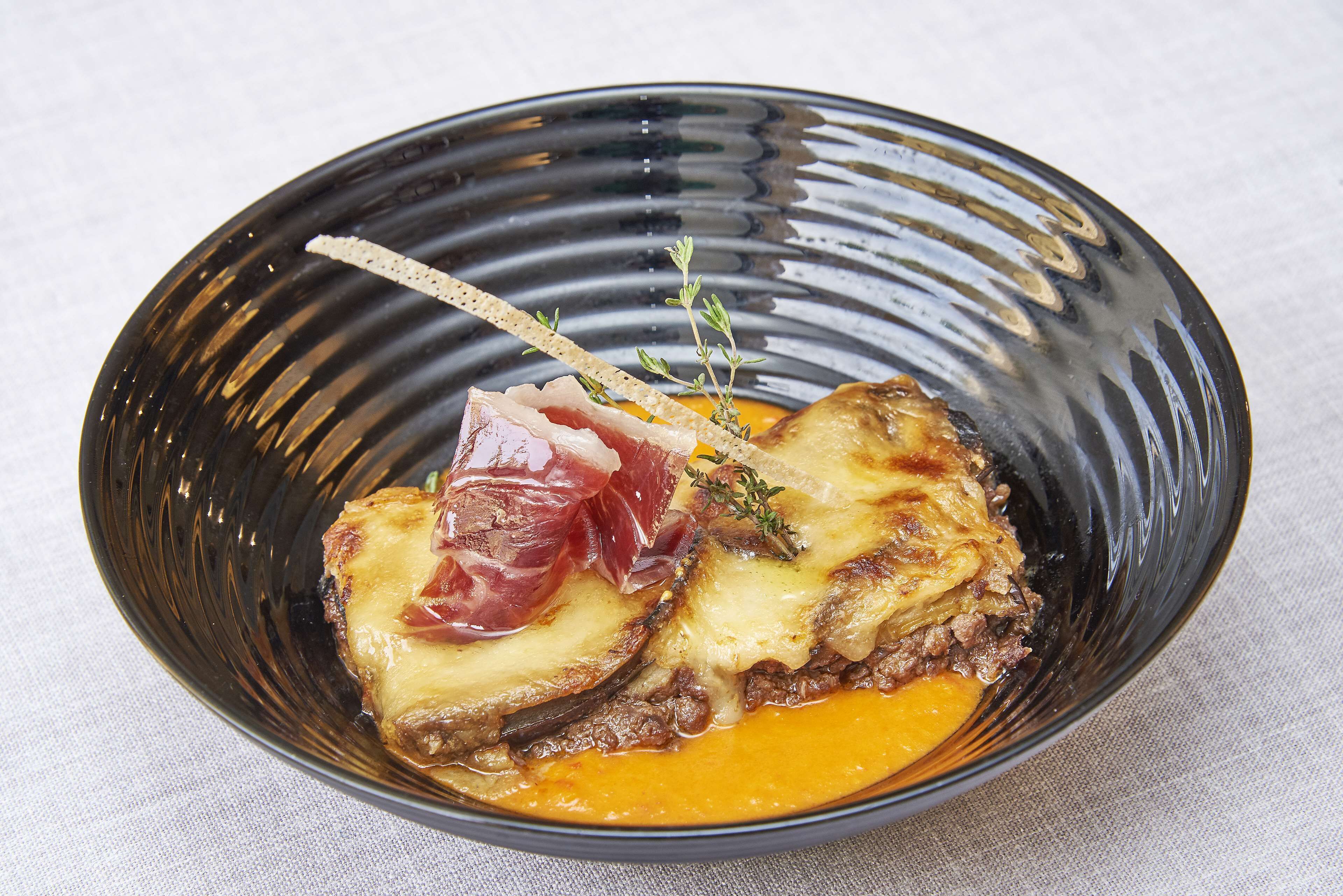 Un tazón negro con un trozo de carne, queso y una guarnición sobre una superficie blanca.