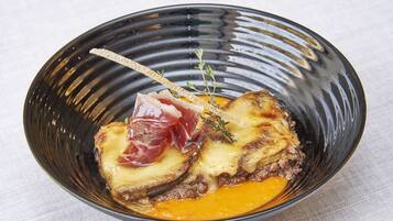 Un tazón negro con un trozo de carne, queso y una guarnición sobre una superficie blanca.